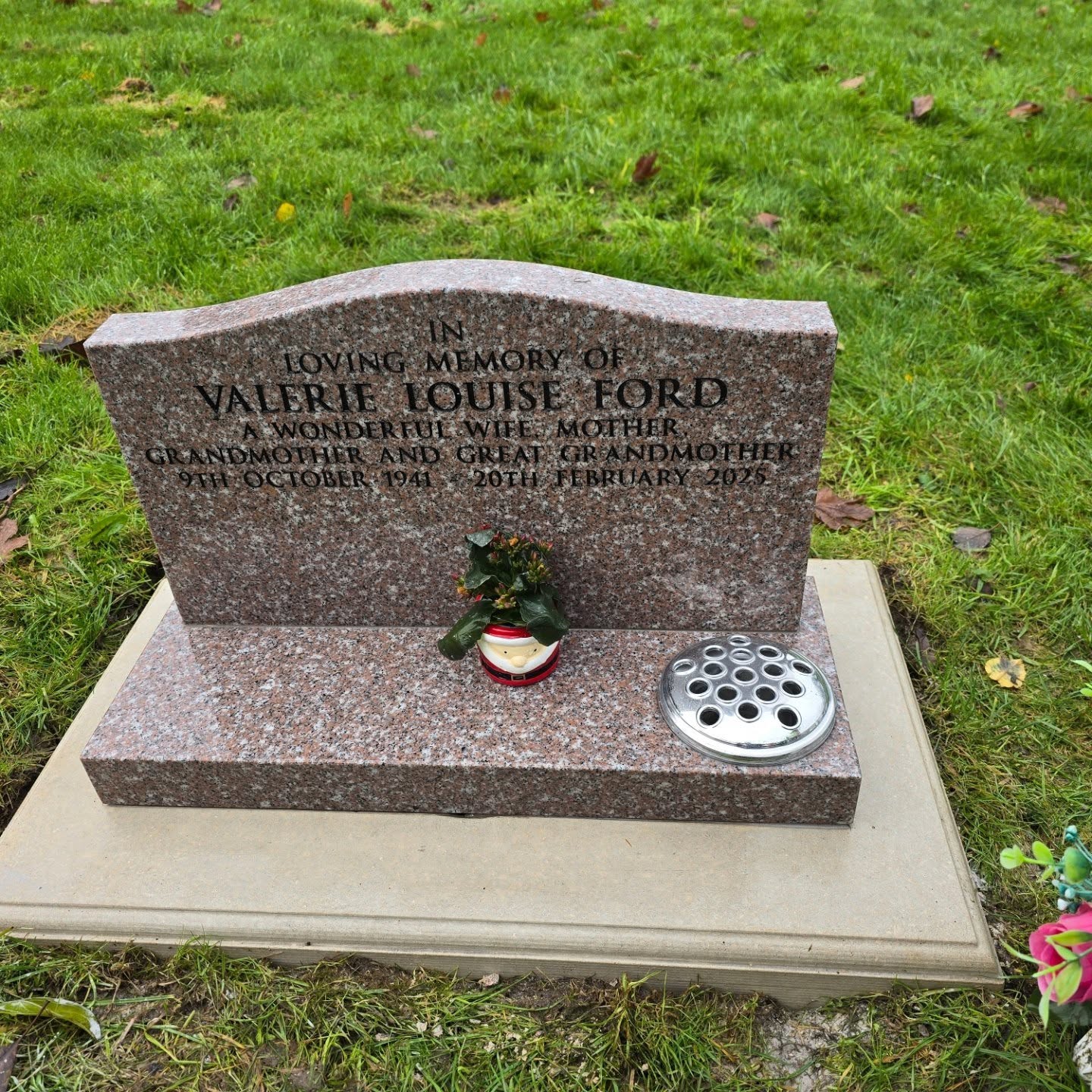 A gravestone on grass reads: "In loving memory of Valerie Louise Ford, a wonderful wife, mother, grandmother, and great grandmother, 1948–2023." A small floral arrangement sits in front.