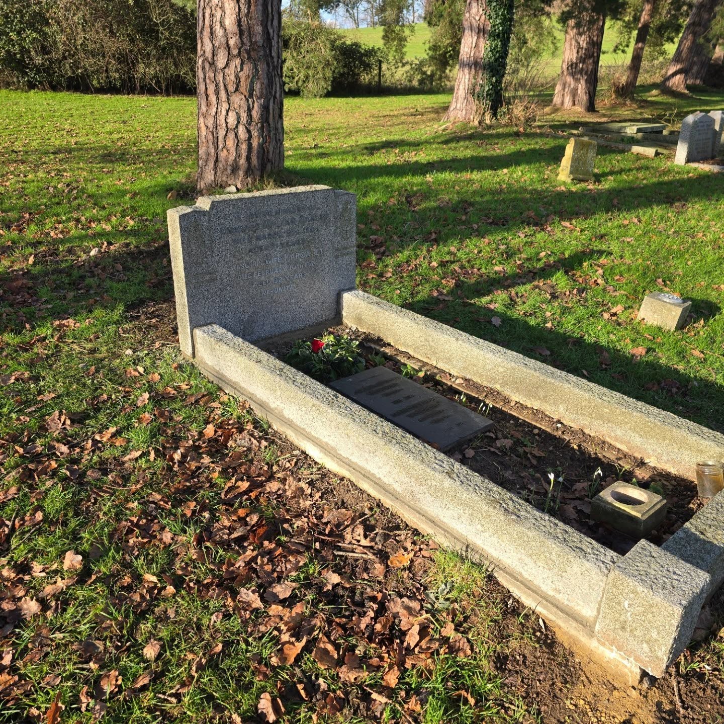 A gravestone and grave plot in a grassy cemetery, surrounded by trees and scattered fallen leaves, with sunlight casting shadows on the ground.