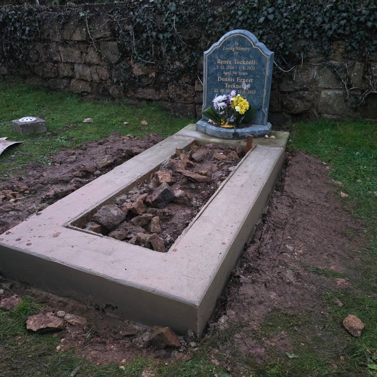 A gravestone with flowers stands at the head of a grave. The grave has fresh concrete edging and loose stones in the center, indicating recent construction or repairs. Grass and a stone wall are in the background.