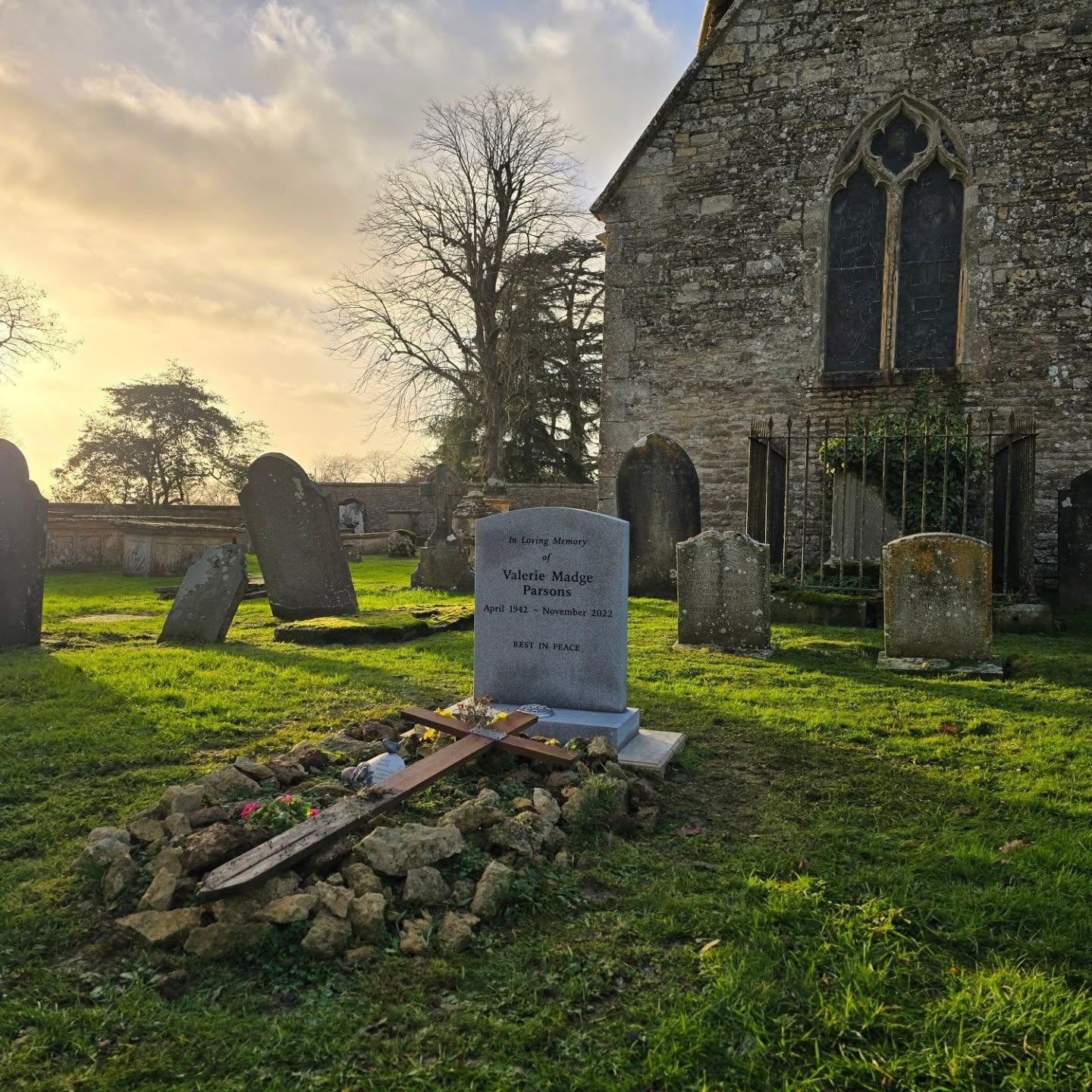 A gravestone with flowers and a wooden cross lies in a grassy cemetery beside an old stone church under a partly cloudy sky at sunset.