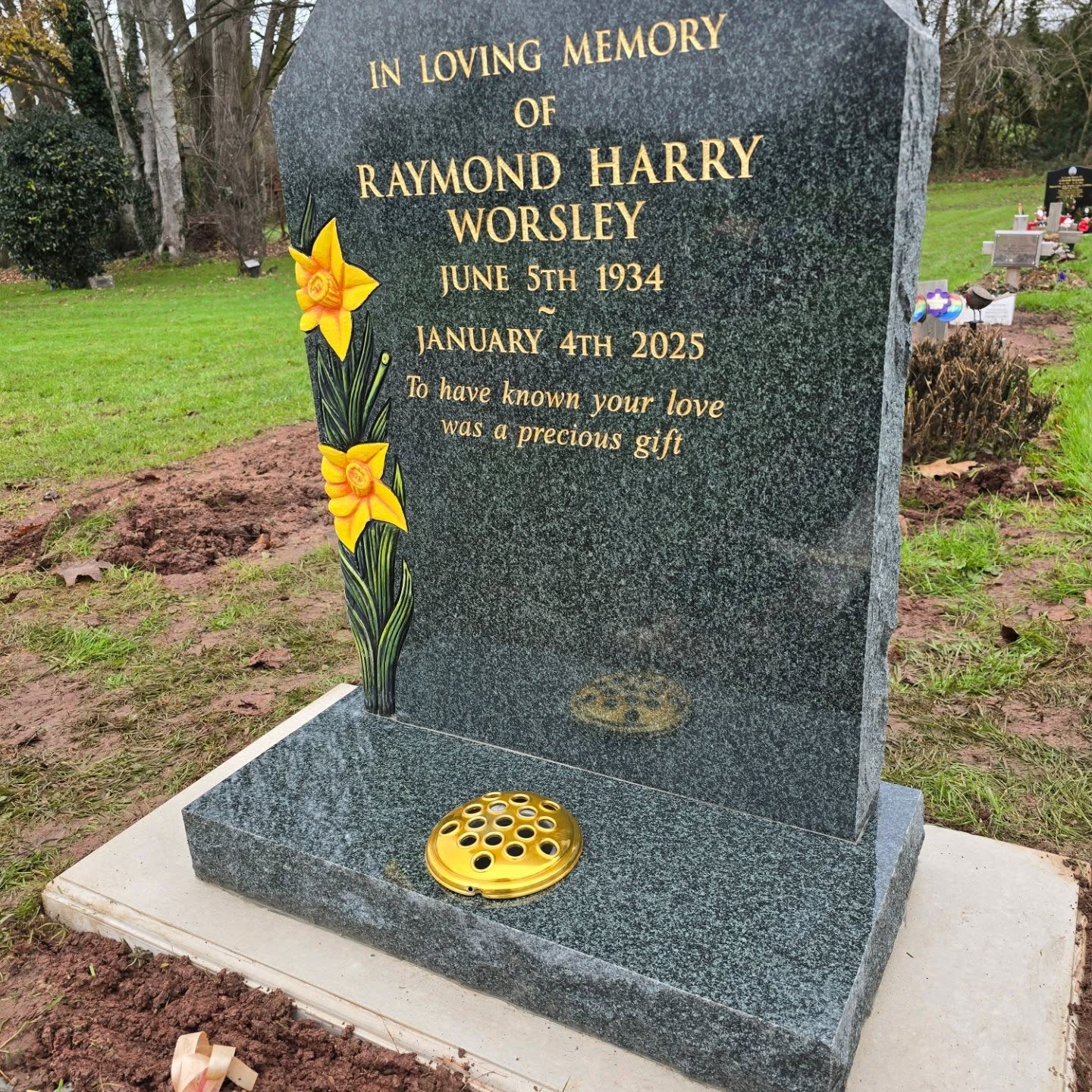 A dark granite headstone with yellow daffodils and gold engraved text memorializing Raymond Harry Worsley, 1934–2025, with the words, "To have known your love was a precious gift.