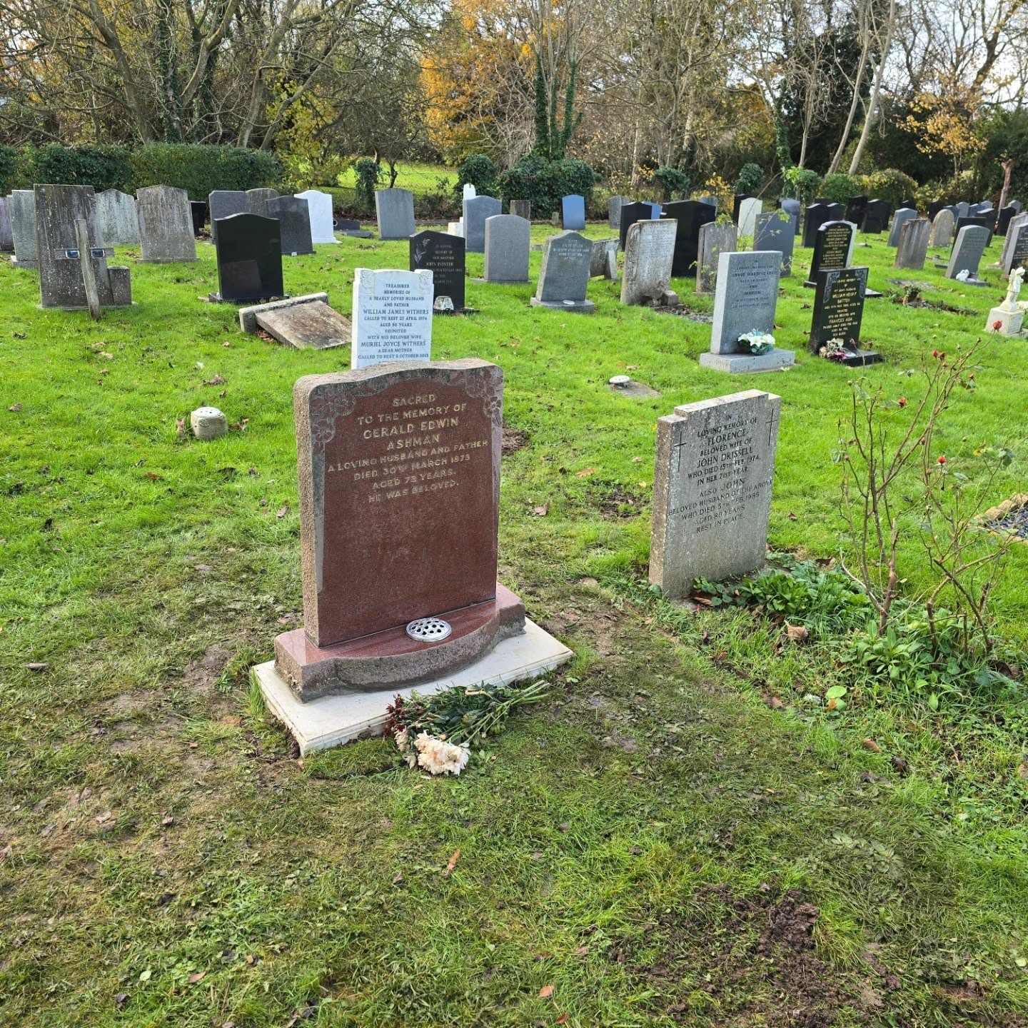 A cemetery with various gravestones on green grass; a prominent red-brown headstone in the foreground and more headstones scattered in the background with some autumn trees visible.