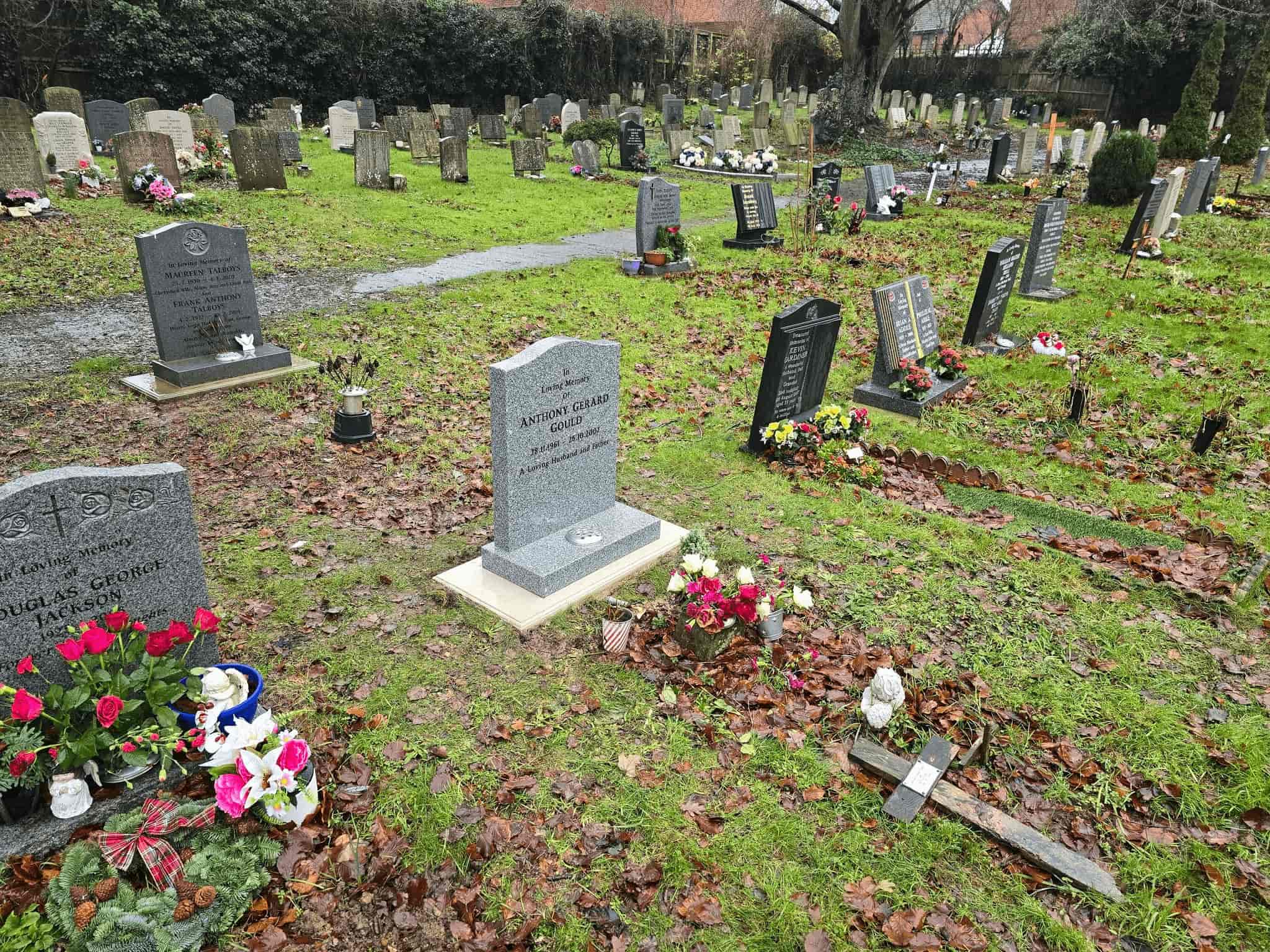 A cemetery with several gravestones and fresh flowers placed on the graves. Fallen leaves scatter the ground, and trees and shrubs line the background on a cloudy day.