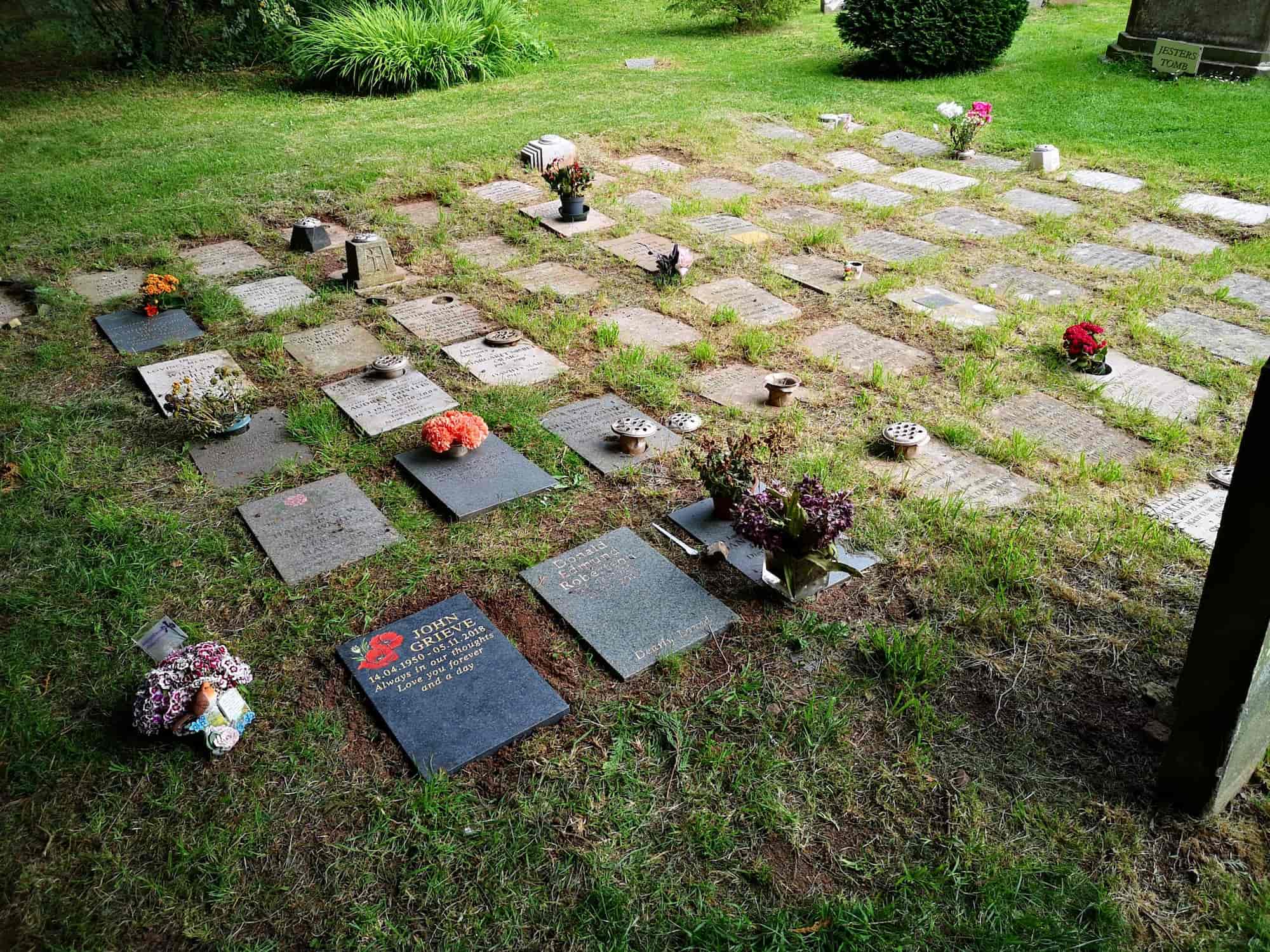 A grassy cemetery with flat stone grave markers arranged in rows, some with flowers and vases placed on top, and small bushes in the background.