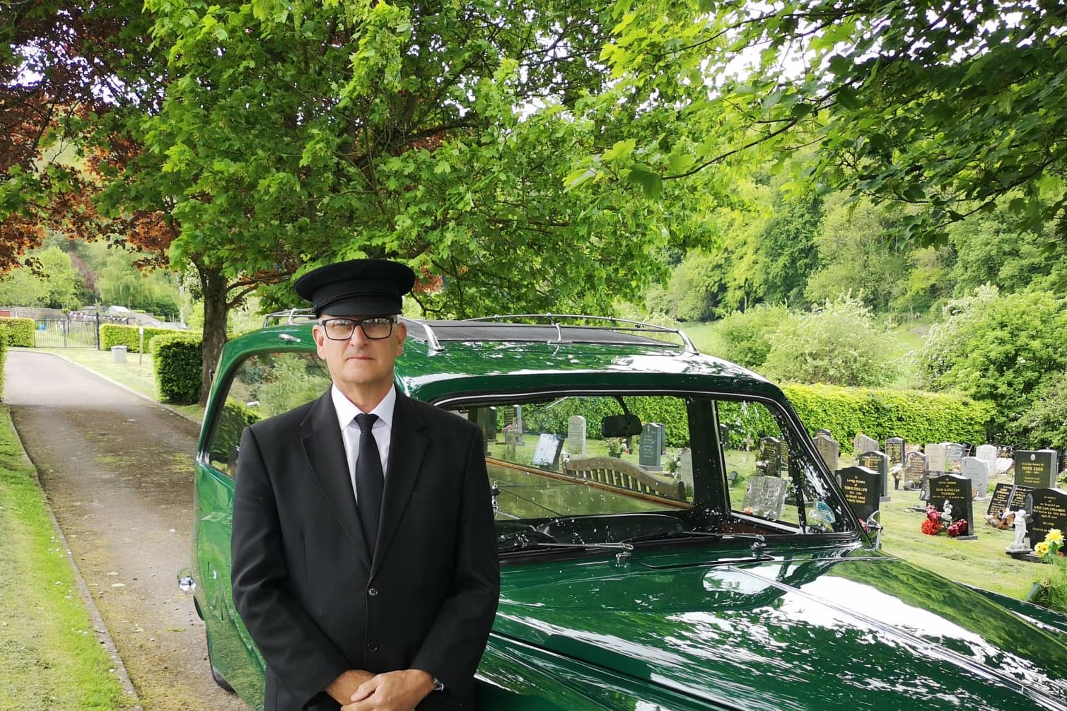 A man in a black suit and hat stands in front of a vintage green hearse, with a cemetery and gravestones in the background, under leafy trees.