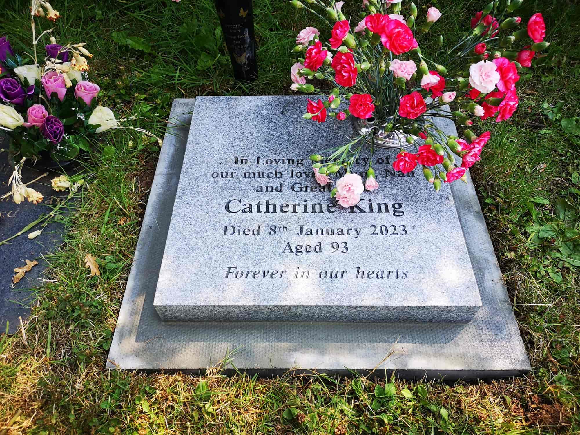 A gravestone for Catherine King, who died January 8, 2023, at age 93. Flowers are placed on and beside the stone, which reads "Forever in our hearts." The grave is surrounded by grass.