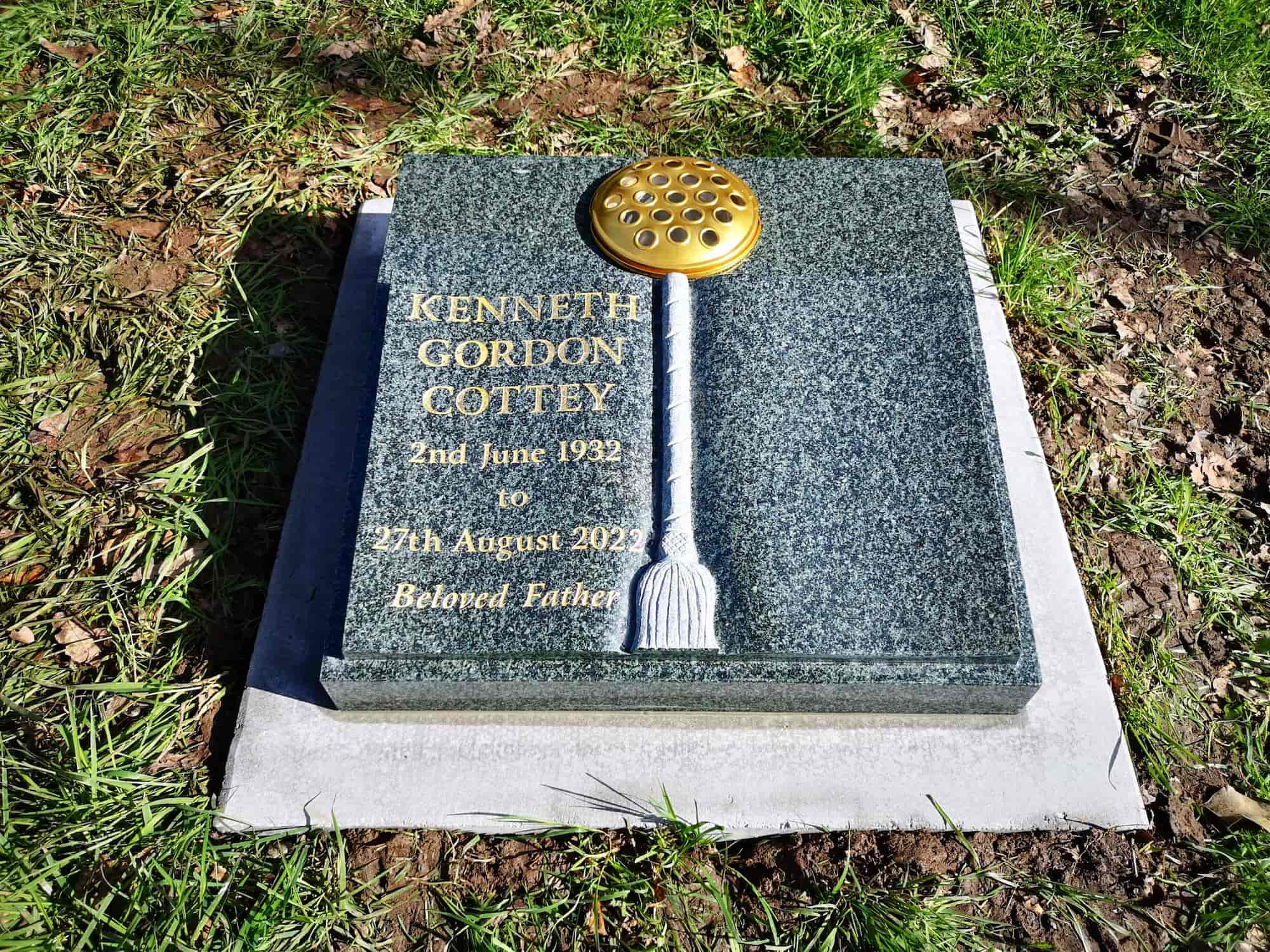 A gravestone for Kenneth Gordon Cottey, 1932–2022, labeled "Beloved Father," with a gold-colored ornament resembling a showerhead and broom handle mounted vertically on the stone. Grass and earth surround the grave.