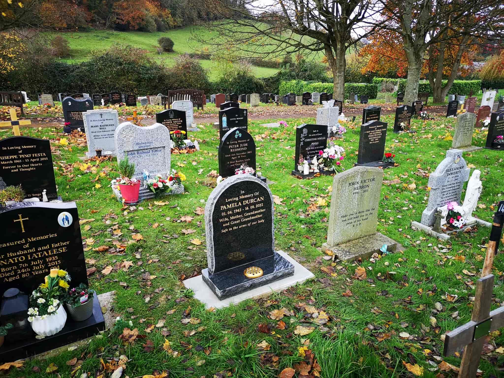 A cemetery with several gravestones, some adorned with flowers and wreaths. Trees with autumn leaves stand in the background, and fallen leaves are scattered on the grass.
